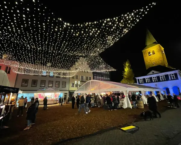 Glitzernde Lichterketten schmücken den **Weihnachtsmarkt in Dorsten**. Menschen bummeln unter Zelten und vor Buden, während im Hintergrund die beleuchtete Kirche thront.