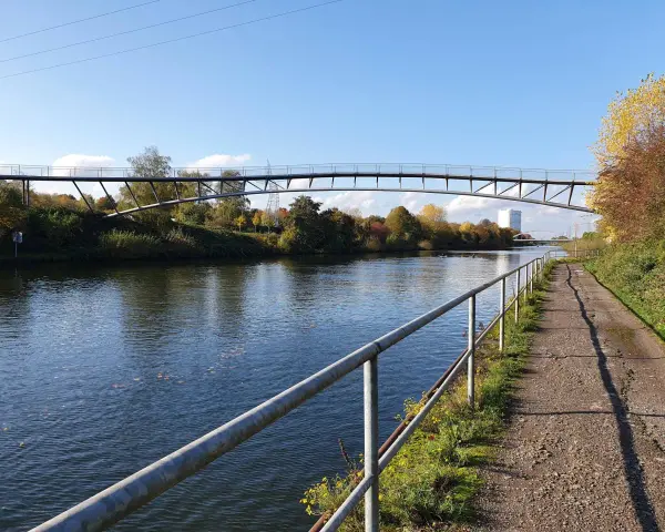 Ein Radweg am Rhein-Herne-Kanal führt unter einer modernen Brücke hindurch. Herbstlaub schmückt die Ufer.