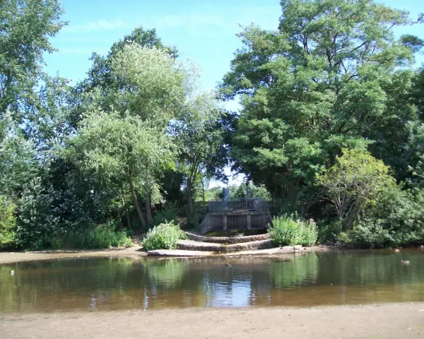 Ein ruhiger blauer See in Dorsten, umgeben von üppigem Grün. Eine kleine Holzbrücke überspannt das Wasser, während Enten friedlich schwimmen.