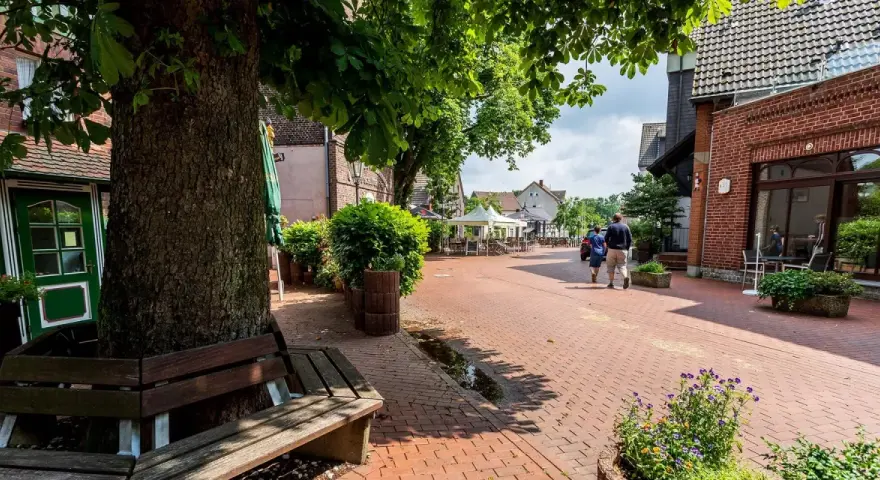 Ein lauschiger Kirchplatz in Recklinghausen mit Kopfsteinpflaster, Bäumen und Sitzgelegenheiten. Ein Paar spaziert vorbei an Cafés mit Außenbestuhlung.