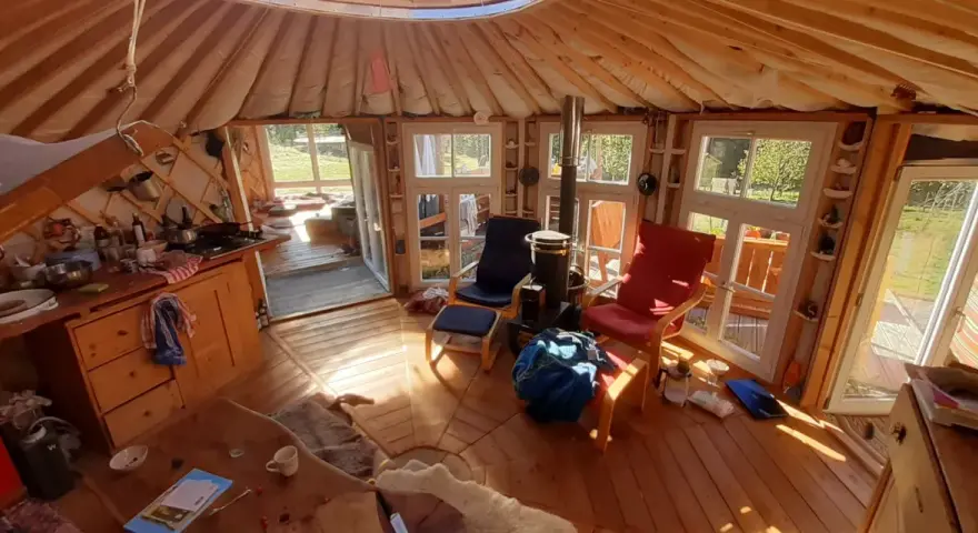 Gemütliches Inneres einer Jurte als Wohnhaus mit Holzofen, Küche und Sitzgelegenheiten. Sonnenlicht fällt durch die Fenster.