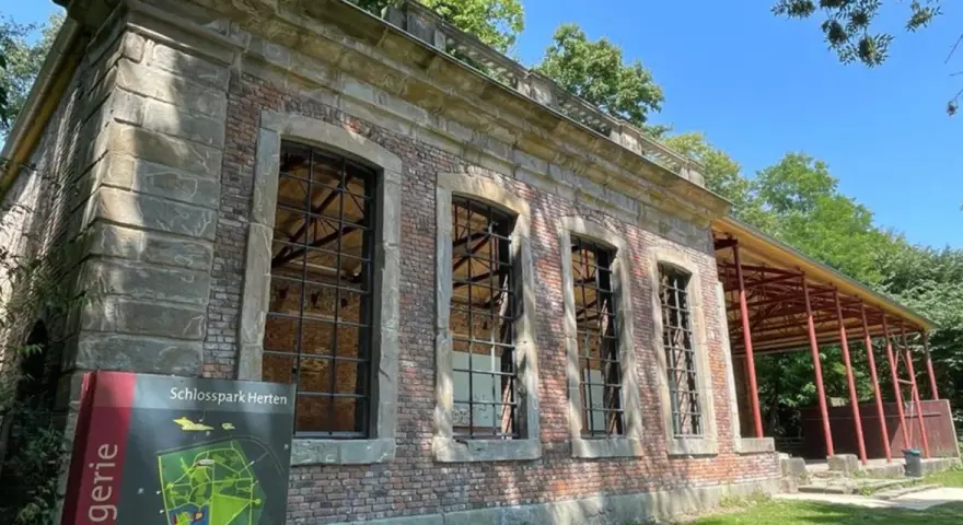 Die Orangerie Herten, ein historisches Gebäude mit großen Fensterfronten, steht im Schatten von Bäumen.