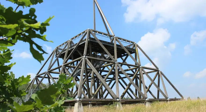 Der Spurwerkturm Waltrop, eine beeindruckende Holzkonstruktion, ragt gegen einen blauen Himmel mit Wolken empor. Grüne Blätter rahmen die Szene ein.