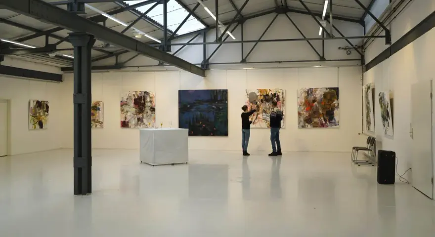 Zwei Personen betrachten Kunstwerke in der Kunsthalle Burkamp. Abstrakte Gemälde hängen an den weißen Wänden, ein Tisch mit Gläsern steht in der Mitte.