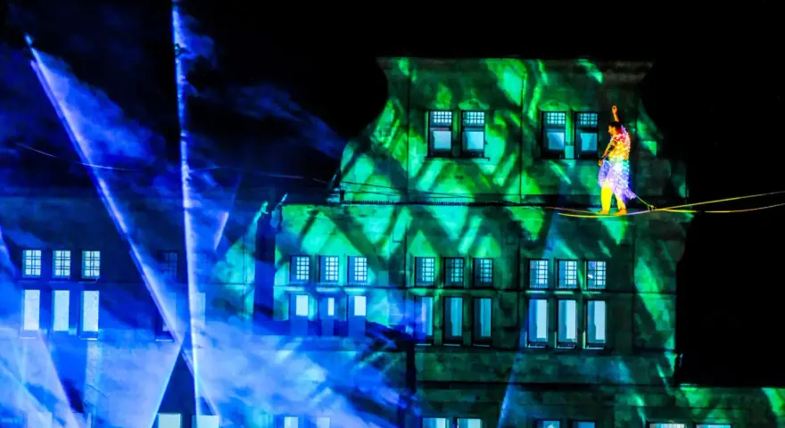 Ein Artist balanciert auf einem Hochseil vor einem Gebäude, das mit grünen Lichtmustern beleuchtet ist. Blaue Lichtstrahlen durchziehen die Szene. Das ist ein Highlight, wann ist Recklinghausen leuchtet!