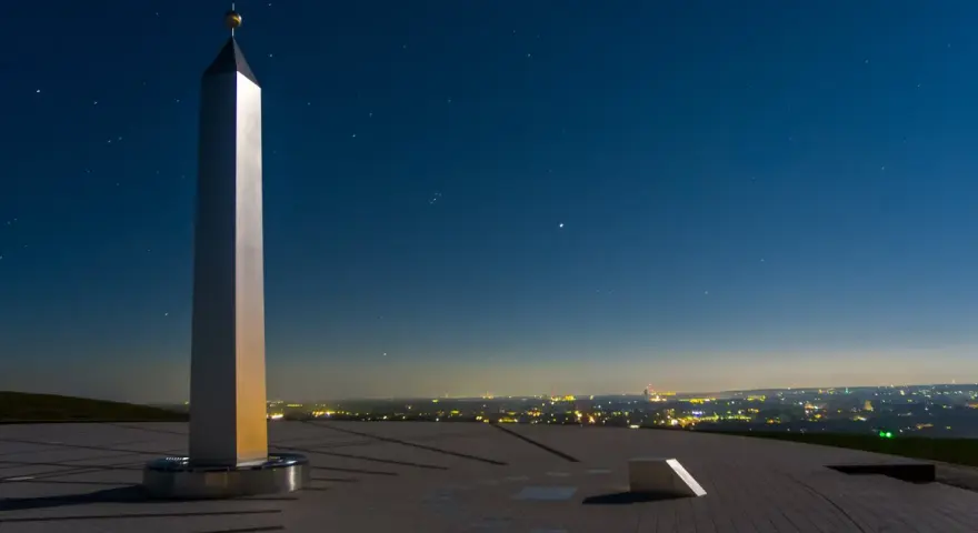 Die Sonnenuhr in Herten ragt unter einem sternenklaren Nachthimmel empor. Ein glänzender Obelisk wirft lange Schatten auf die gepflasterte Fläche.