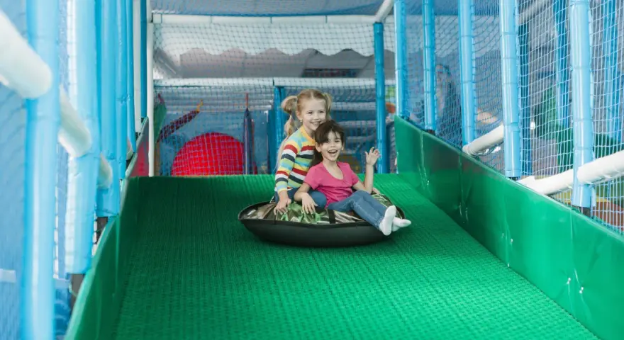 Zwei lachende Mädchen rutschen auf einem Schlauchboot in einem Indoor-Spielplatz.