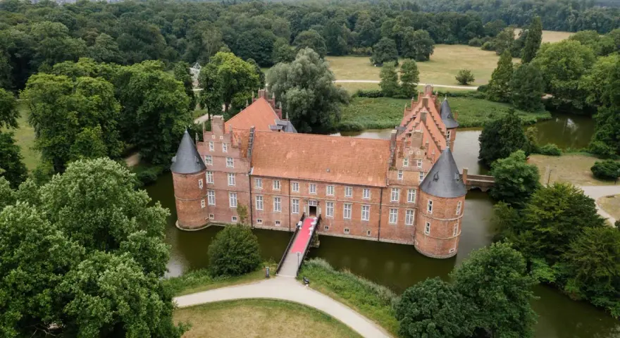 Schloss Recklinghausen, ein imposantes Wasserschloss, umgeben von grünen Bäumen und einem Teich. Eine rote Teppich führt zur Eingangstür.