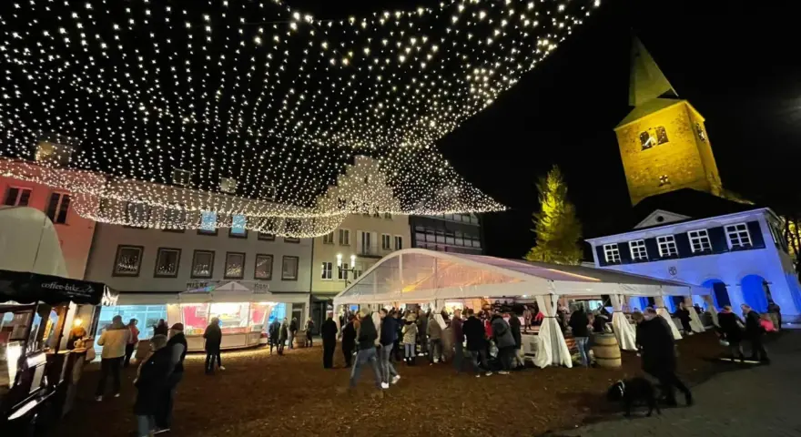 Glitzernde Lichterketten schmücken den **Weihnachtsmarkt in Dorsten**. Menschen bummeln unter Zelten und vor Buden, während im Hintergrund die beleuchtete Kirche thront.