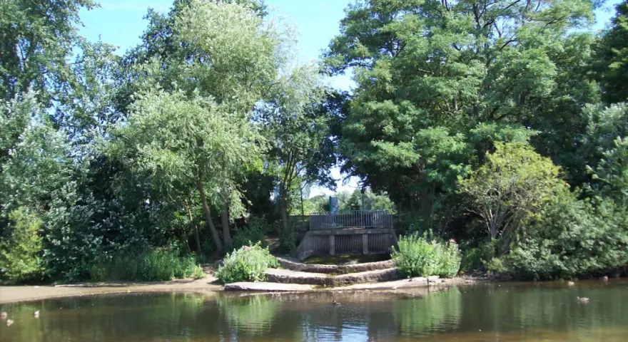 Ein ruhiger blauer See in Dorsten, umgeben von üppigem Grün. Eine kleine Holzbrücke überspannt das Wasser, während Enten friedlich schwimmen.