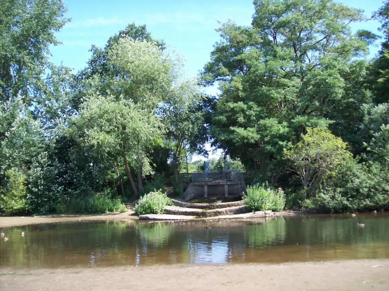 Idyllischer blauer See in Dorsten, umgeben von üppigem Grün. Eine kleine Brücke überspannt das Wasser, während Enten friedlich schwimmen.