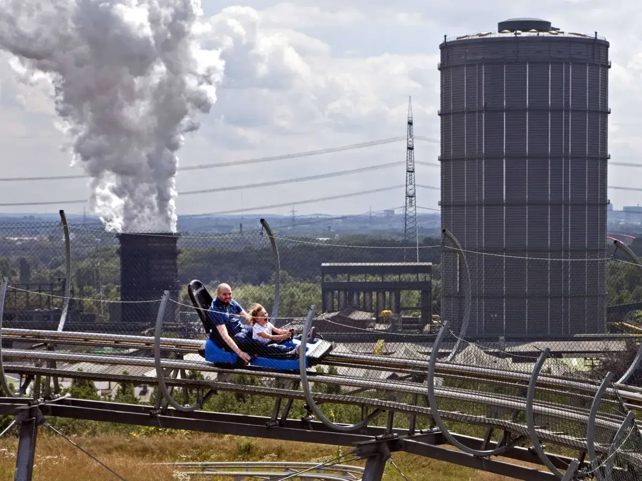 Vater und Kind auf der Sommerrodelbahn Bottrop, mit rauchendem Schornstein und Gasometer im Hintergrund.