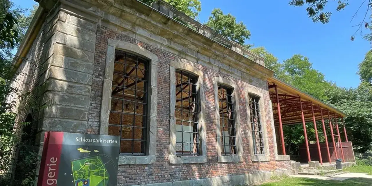 Die Orangerie Herten, ein historisches Gebäude mit großen Fensterfronten und einem roten Vordach, steht im Schatten von Bäumen.