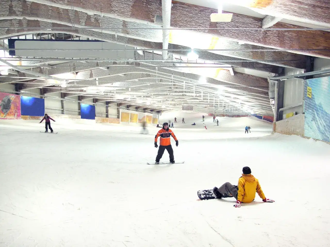 Schneebedeckte Piste in der Bottroper Skihalle. Ein Snowboarder in Orange steht, ein anderer sitzt im gelben Anzug.
