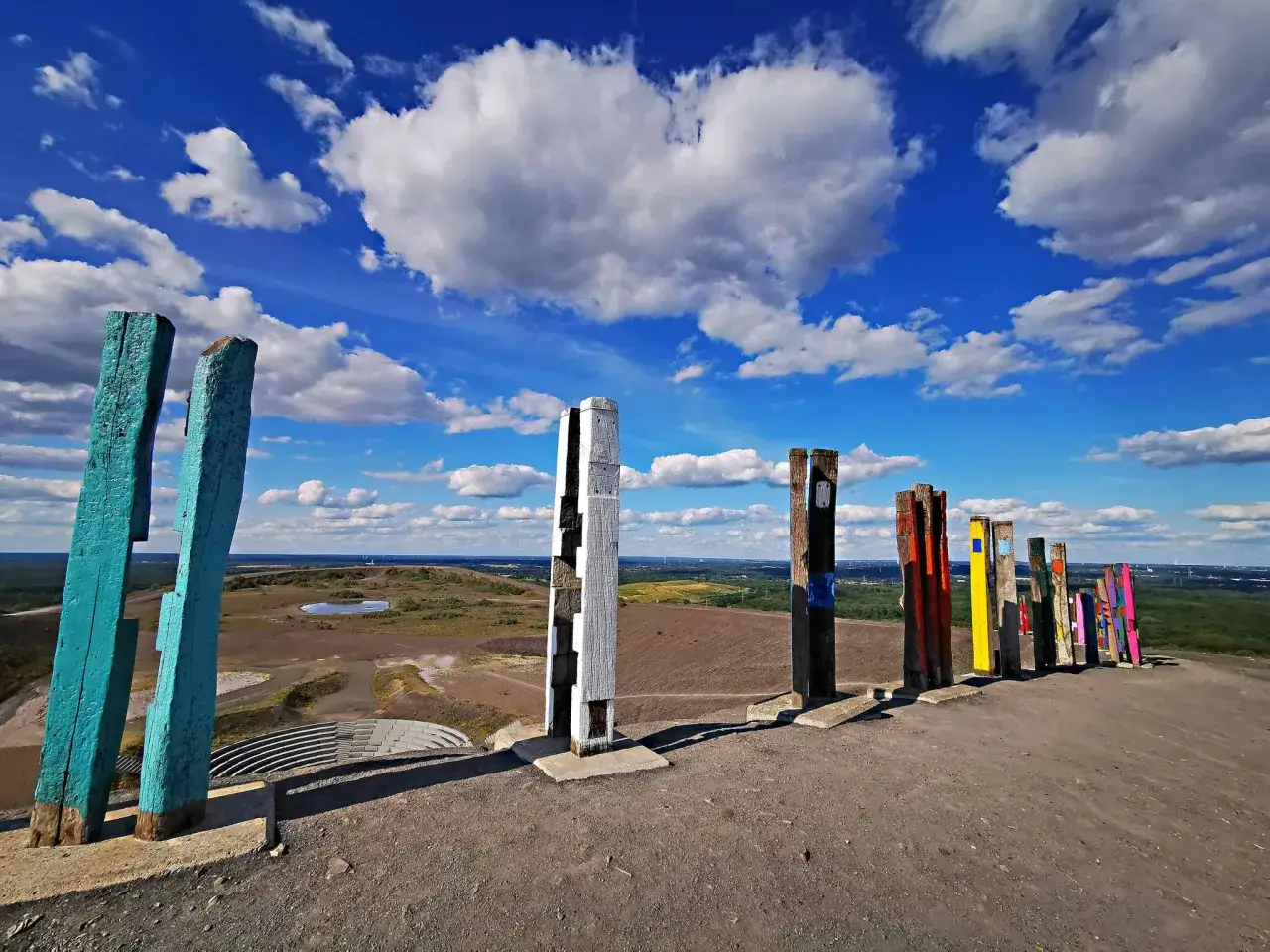 Bunte Holzskulpturen säumen den Weg auf der Halde Haniel unter einem strahlend blauen Himmel mit weißen Wolken.