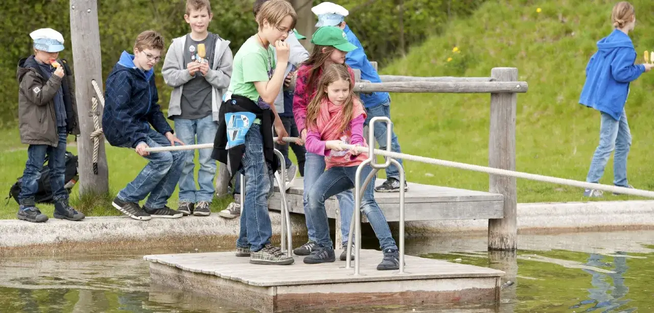 Kinder spielen am Schiffshebewerk Henrichenburg Spielplatz. Sie ziehen an einem Seil, um eine Holzplattform zu bewegen.