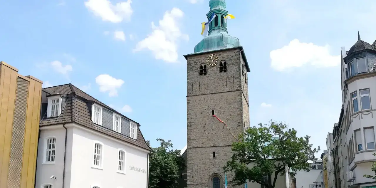 Der Kirchplatz Recklinghausen zeigt einen steinernen Kirchturm mit Uhr und Fahnen unter blauem Himmel. Daneben ein wei&szlig;es Geb&auml;ude.