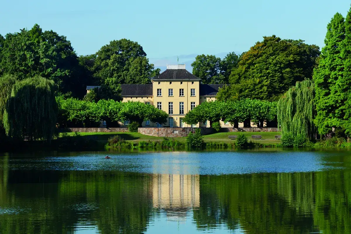 Ein gelbes Herrenhaus mit symmetrischen Fenstern, umgeben von üppigem Grün und einem spiegelnden See. Ein idyllischer Ort zum wohnen in Krefeld.