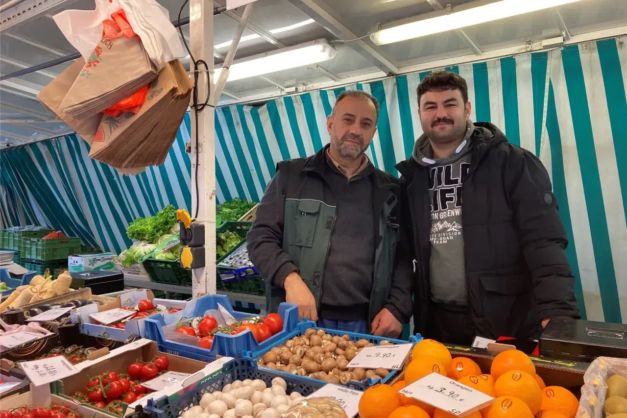 Zwei M&auml;nner stehen an einem Obst- und Gem&uuml;sestand auf dem markt kreis recklinghausen. Frische Tomaten, Pilze und Orangen sind zu sehen.