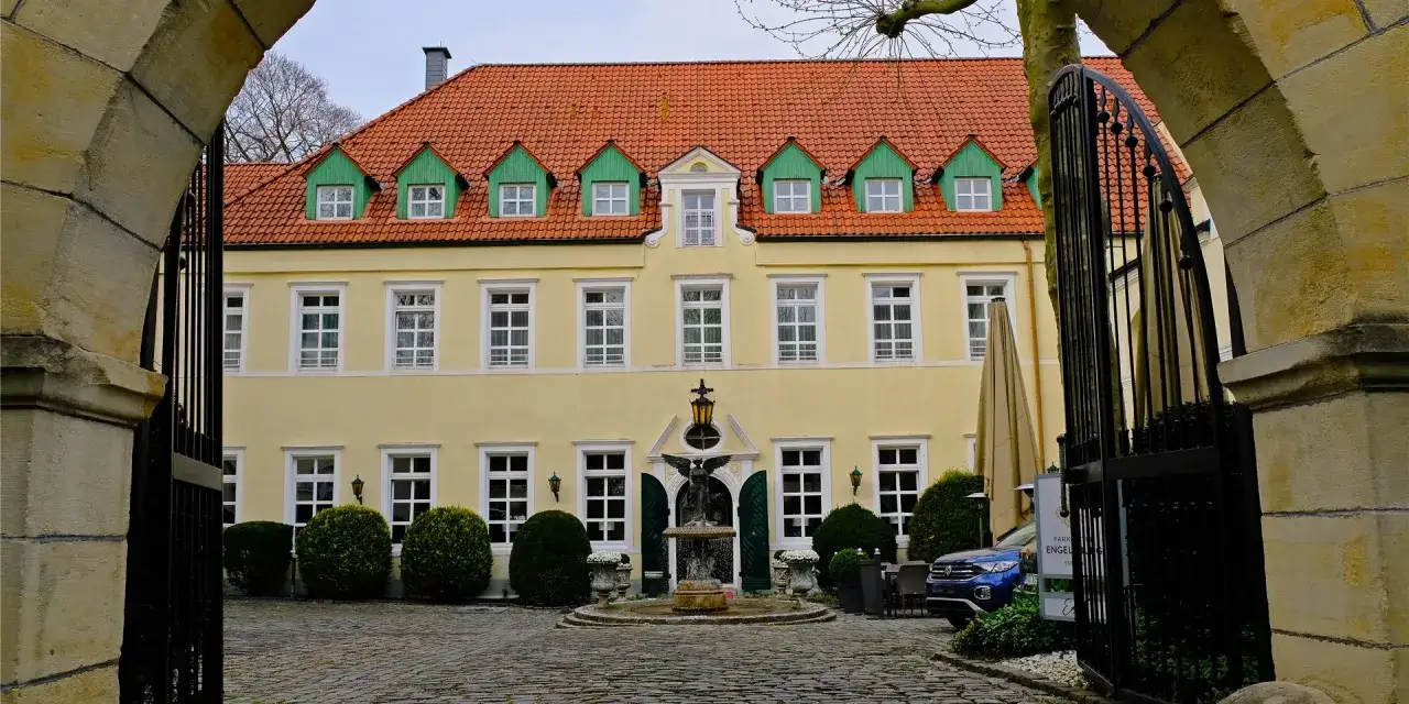 Gelbes Geb&auml;ude mit roten Ziegeldach und gr&uuml;nen Gauben, davor ein Brunnen mit Engelstatue. Das Schloss Recklinghausen l&auml;dt zum Verweilen ein.