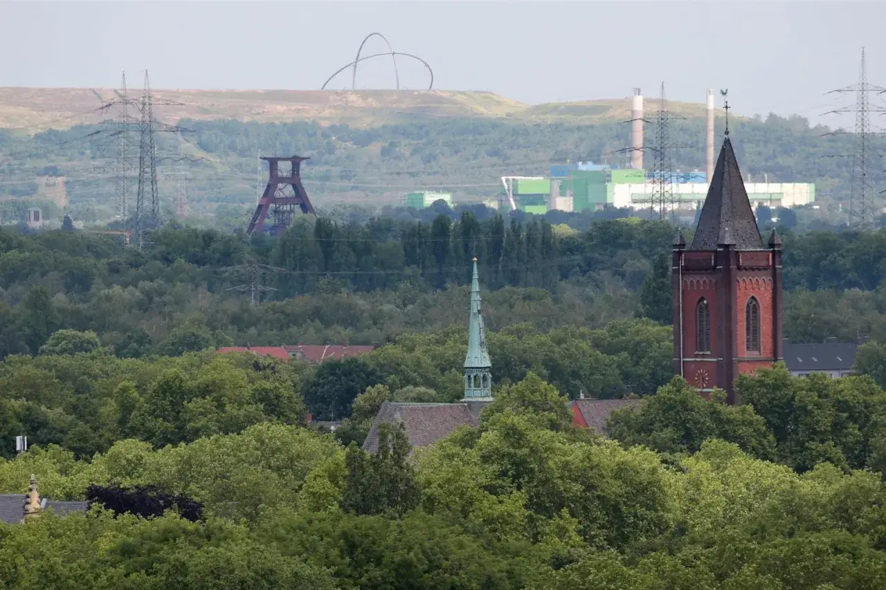 Kirchturm ragt aus B&auml;umen, im Hintergrund die Zeche Ewald Fortsetzung mit F&ouml;rderturm und Industriebauten.