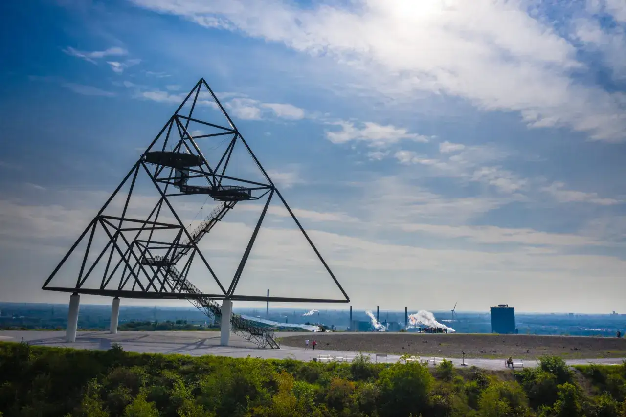 Pyramidenartige Aussichtsplattform auf einer Halde im Ruhrgebiet, mit rauchenden Industrieanlagen im Hintergrund.