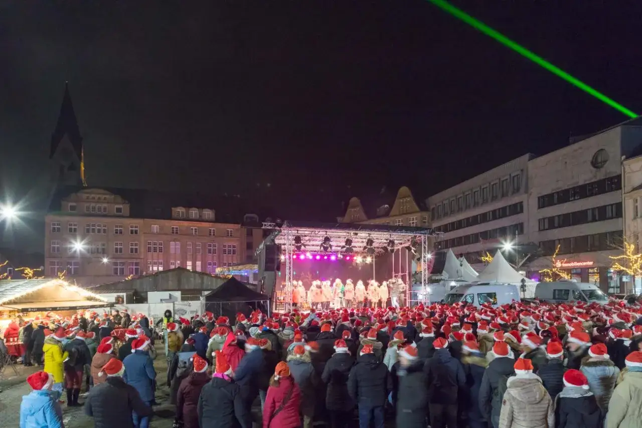 Gro&szlig;e Menschenmenge mit Weihnachtsm&uuml;tzen auf dem Castrop Rauxel Weihnachtsmarkt, w&auml;hrend eine Gruppe auf der B&uuml;hne singt.