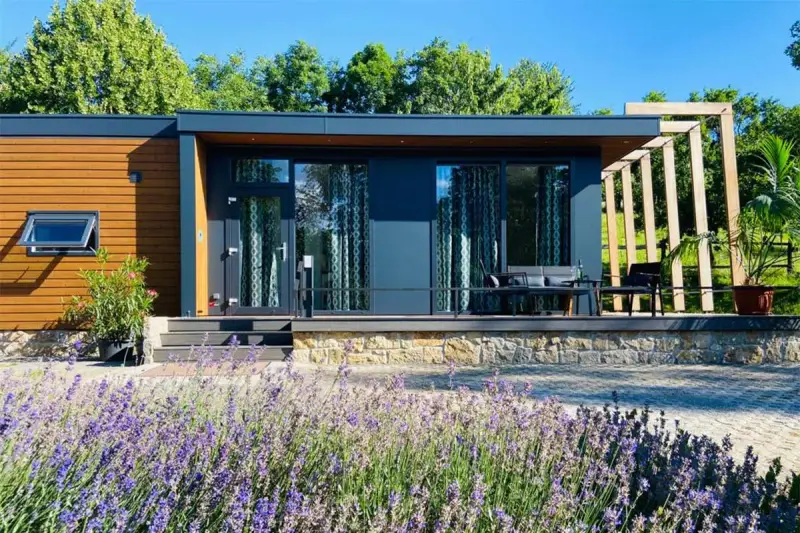 Modernes tiny house im garten der eltern, umgeben von Lavendel und Bäumen. Eine Terrasse mit Sitzmöbeln lädt zum Entspannen ein.